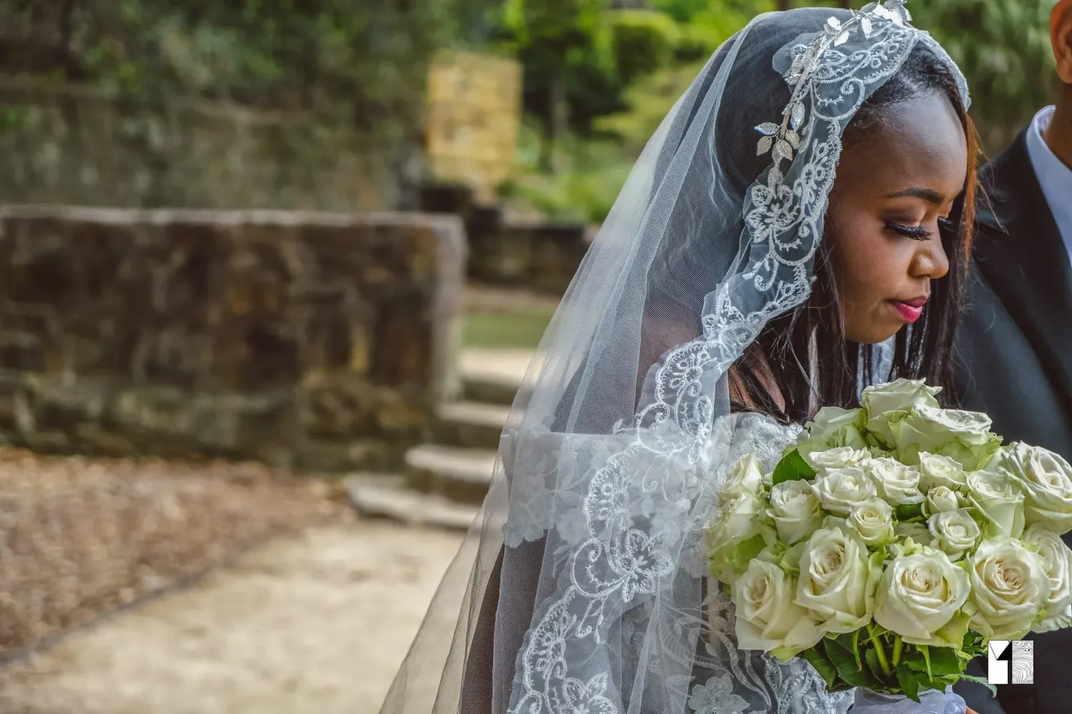 Bride in lace veil looking down at white rose bouquet beside stone steps.