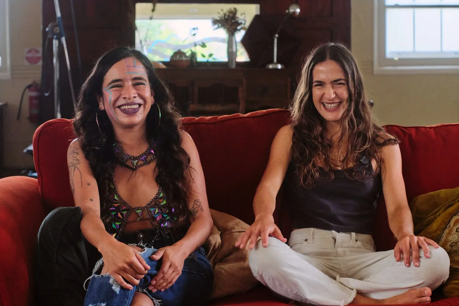 Two smiling women sit together on a red couch in a warmly lit living room setting.