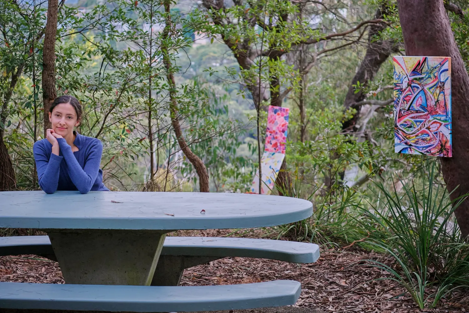 Woman resting at an outdoor picnic table with colorful artwork displayed in Australian bushland.