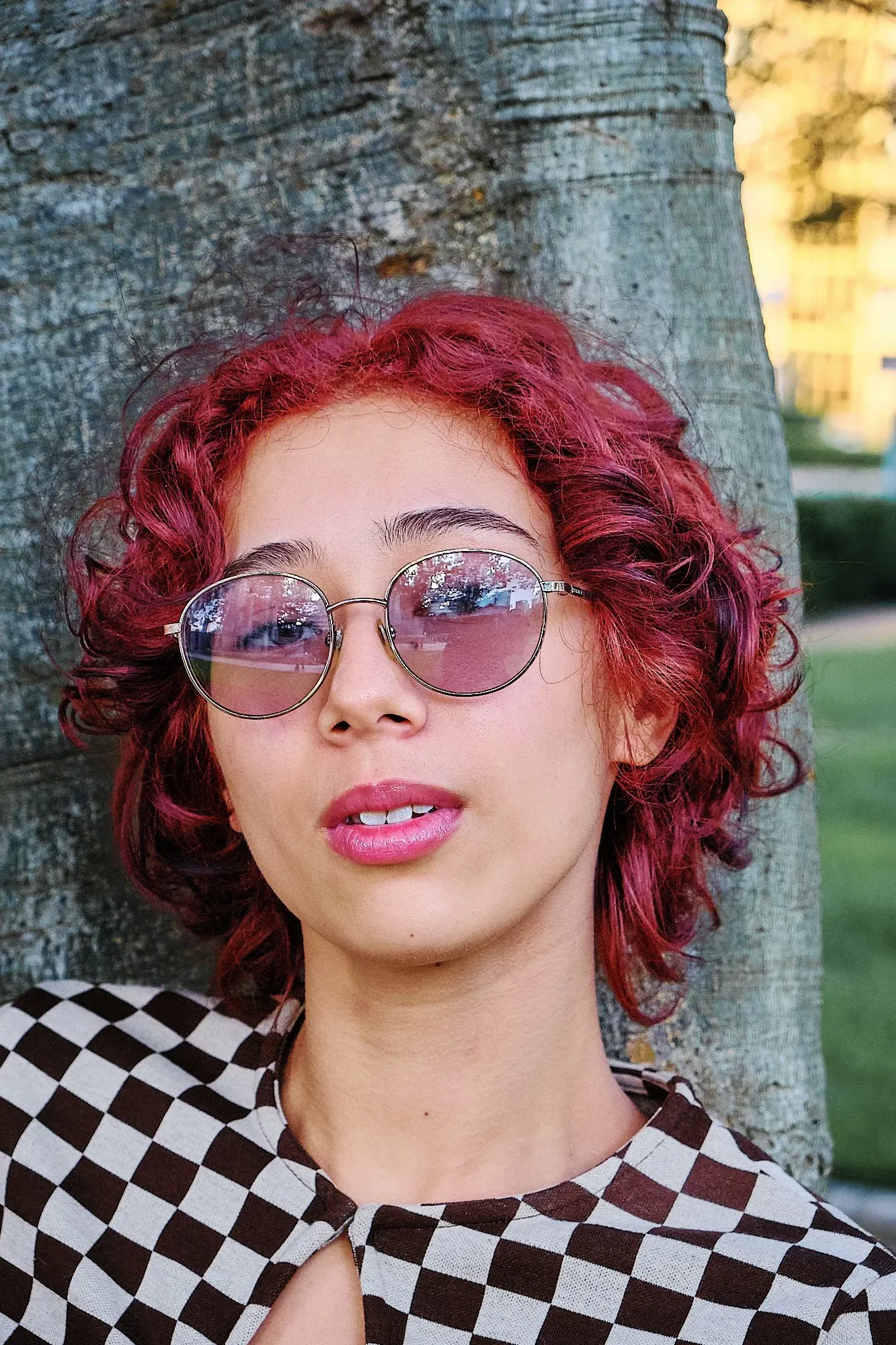 Close-up of woman with vibrant red curly hair wearing decorative round sunglasses by a tree.