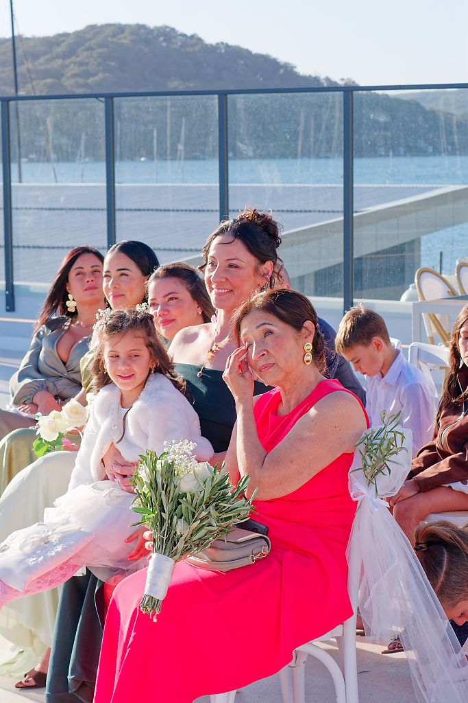 Emotional wedding guests seated outdoors near the water, including a woman in pink wiping a tear and a girl holding a white bouquet.