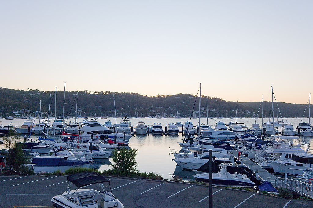 Sailboats and motor yachts moored in a calm marina at dusk with a tree-covered hillside and soft pastel sky in the background.