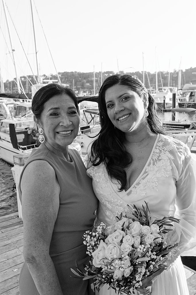Black and white photo of a smiling bride holding a rose bouquet beside a woman in a sleeveless dress at a sunny marina.