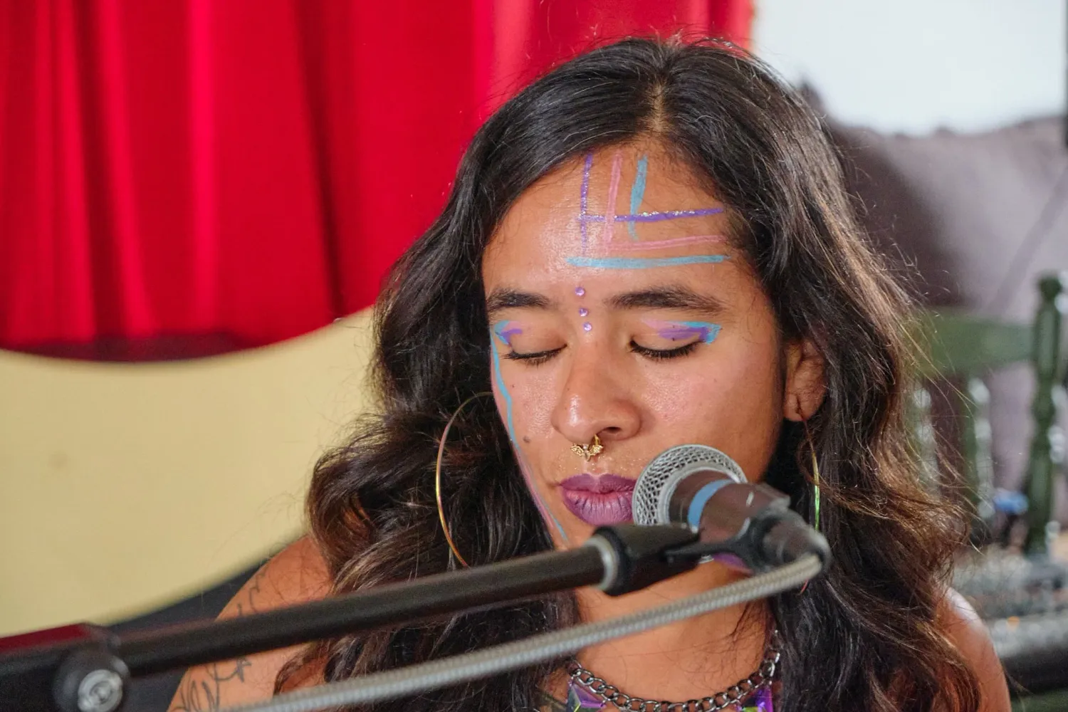 Close-up of female singer at microphone with colorful face paint and purple lipstick, eyes closed.