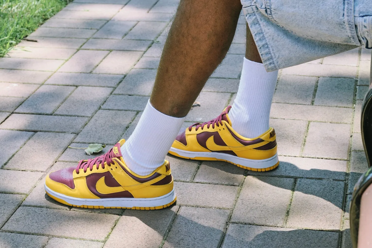 Close-up of yellow and burgundy Nike Dunk Low sneakers worn with white socks on brick pavement.