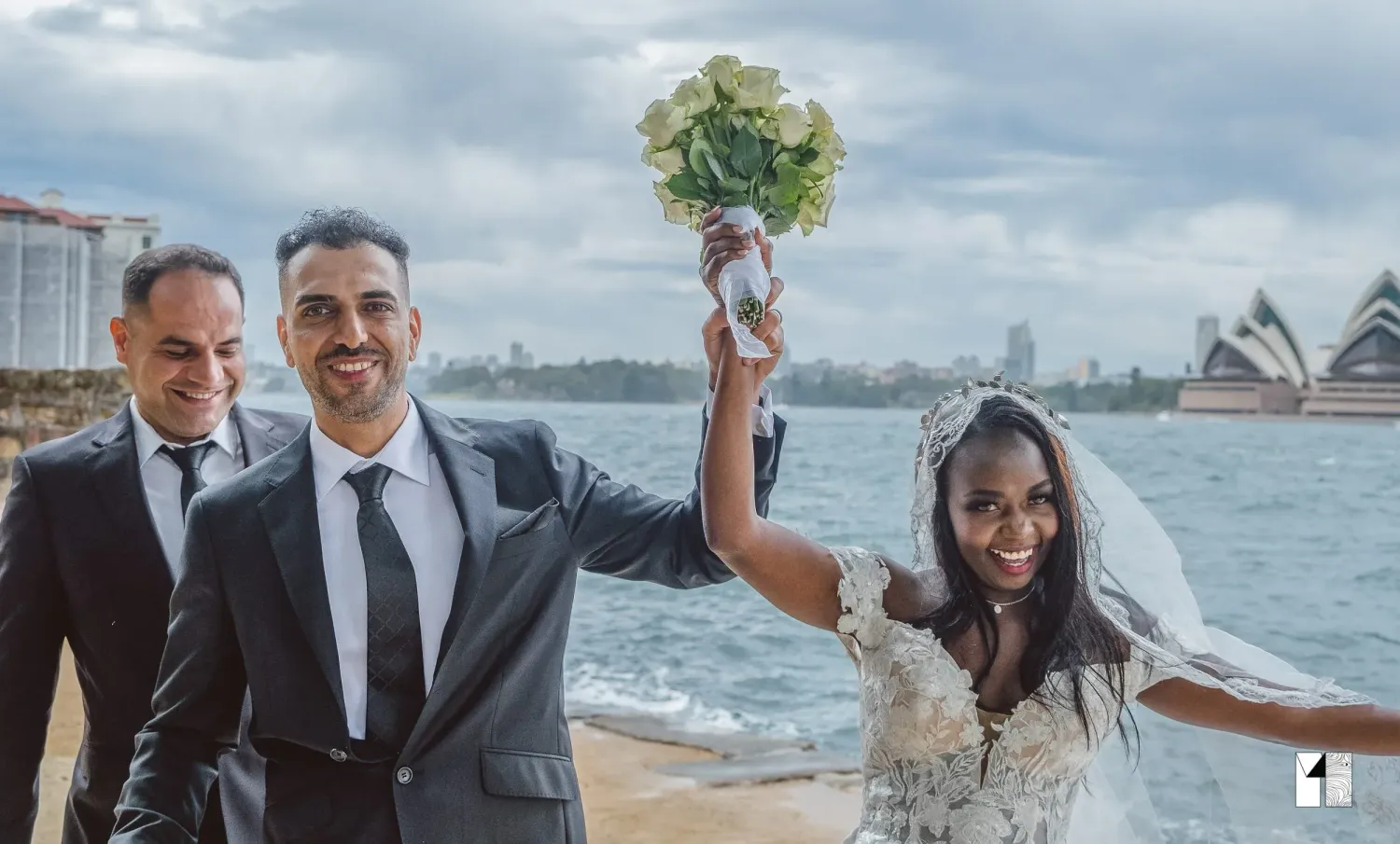 Bride raising bouquet in celebration with groom and guest by Sydney Harbour.
