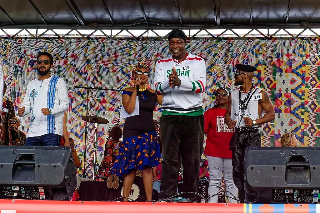 Performers in colorful outfits dance and sing on a vibrant patterned stage at an outdoor festival.