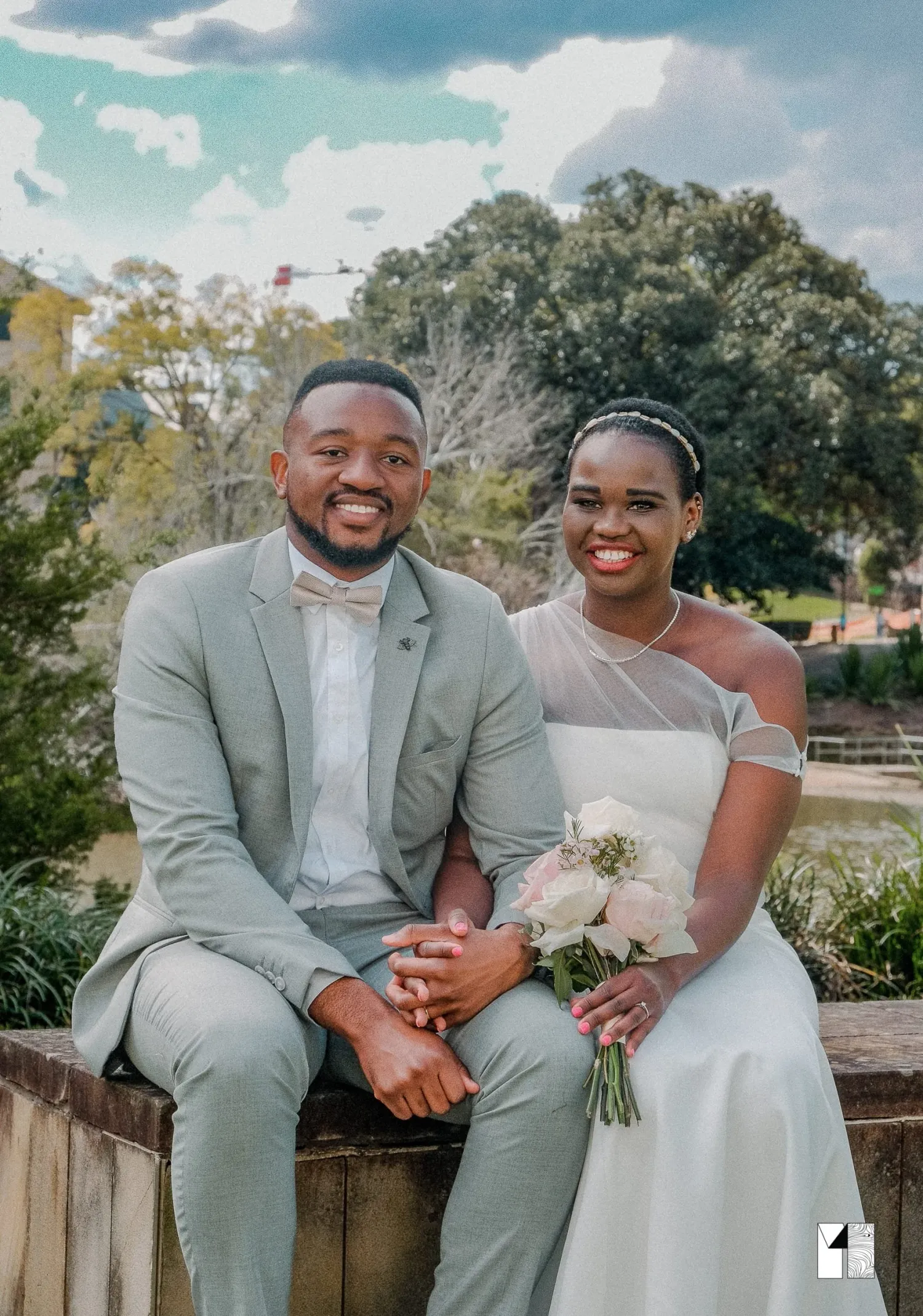 Smiling African couple in wedding attire sitting outdoors, groom in grey suit, bride holding bouquet.