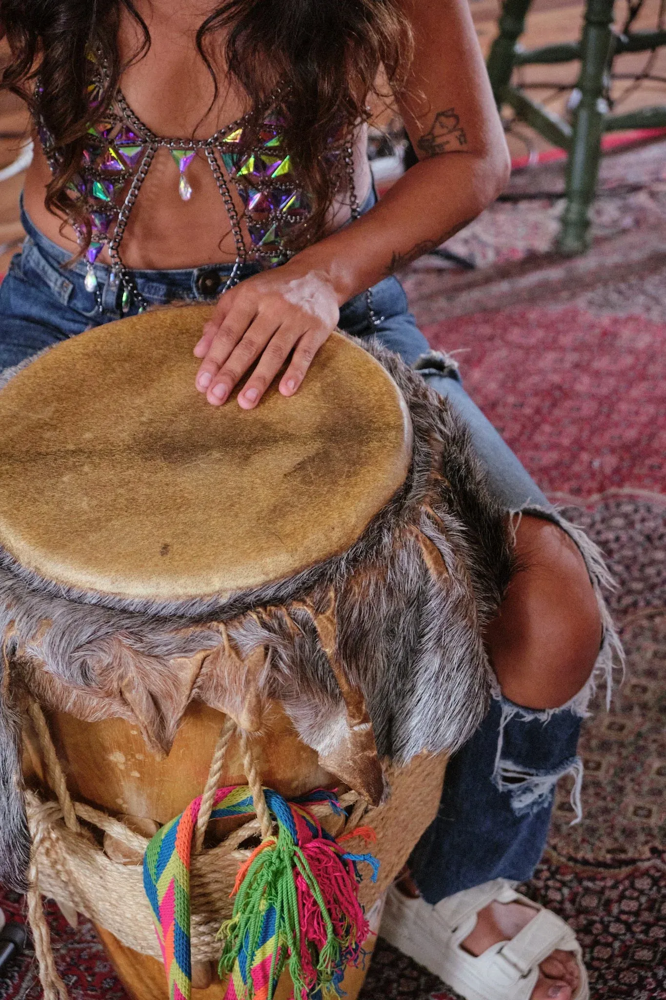 Close-up of woman in embellished top playing a fur-covered djembe drum with colorful tassels.