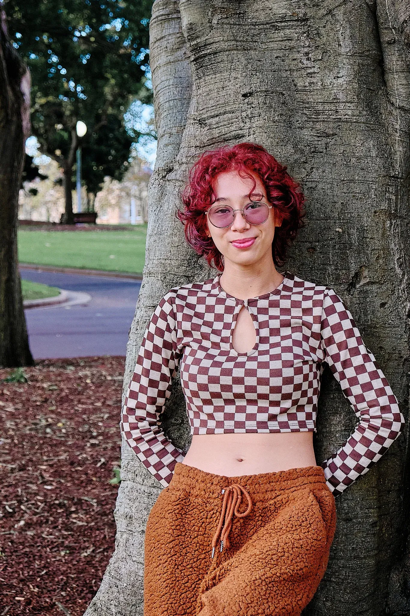 Red-haired woman in checkered crop top and orange pants leaning against large tree in park.