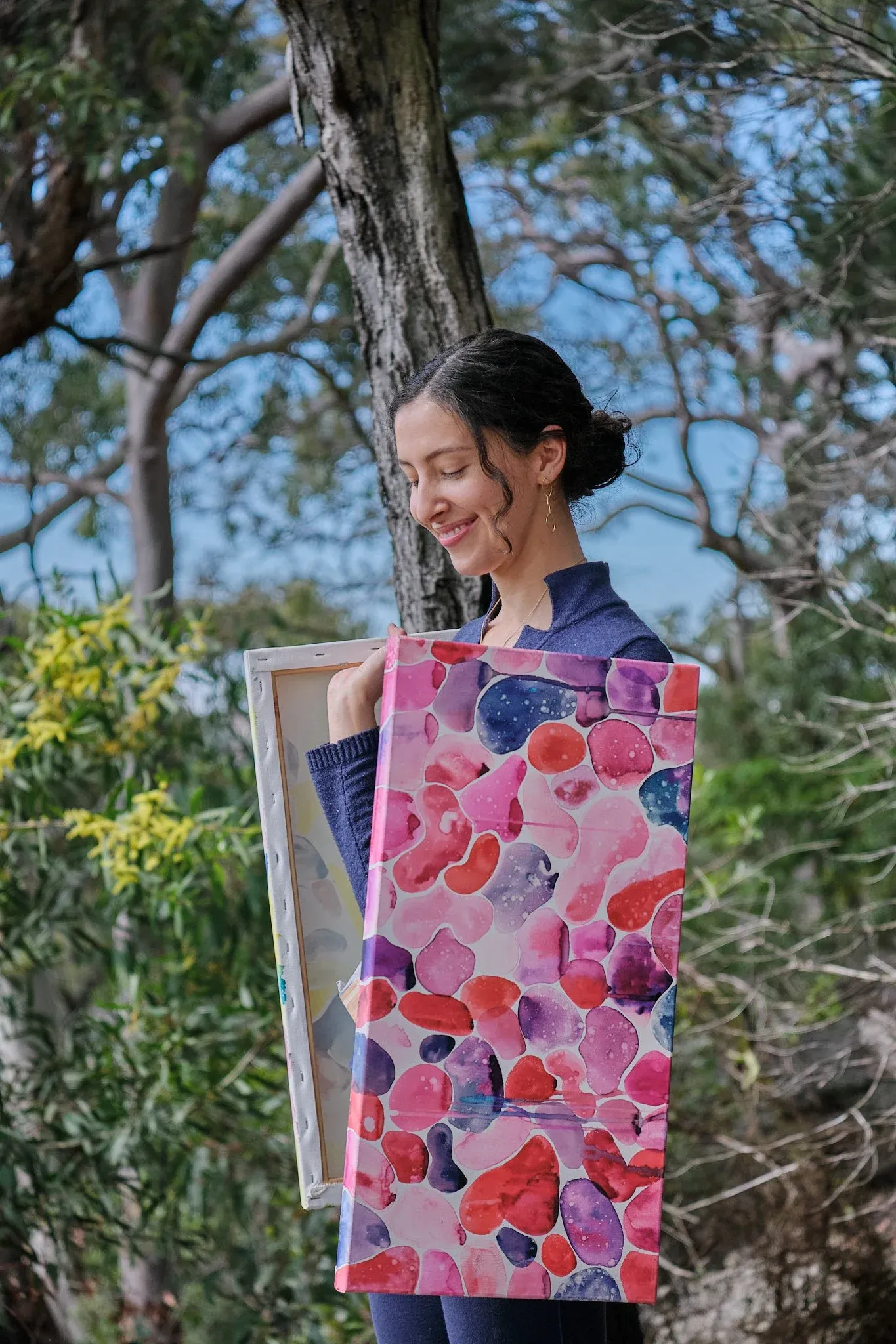 Smiling woman holding a vibrant pink and red abstract canvas painting in Australian bushland.