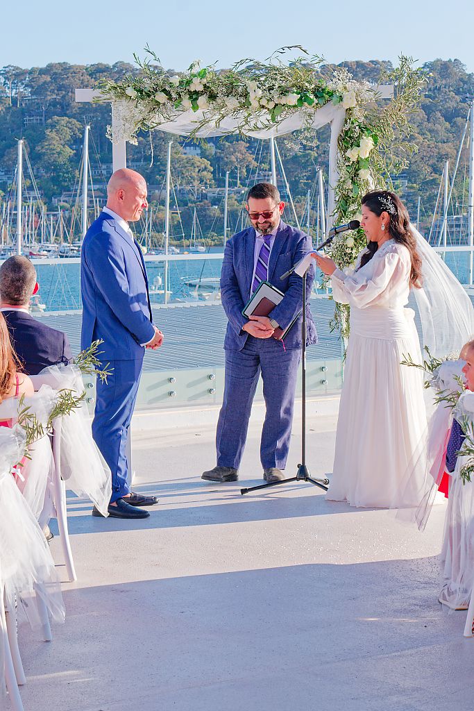Bride in a white gown speaks into a microphone facing the groom in a blue suit during a waterfront wedding ceremony under a floral arch.