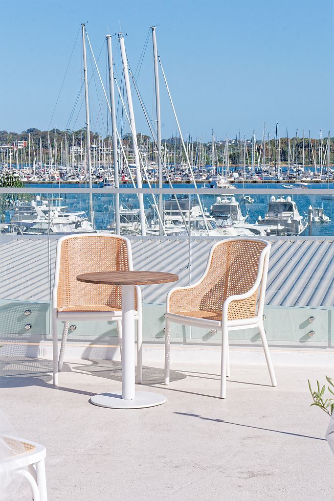 Two white cane-back chairs and a small round wooden table on a sunny waterfront terrace overlooking a marina full of sailboats.