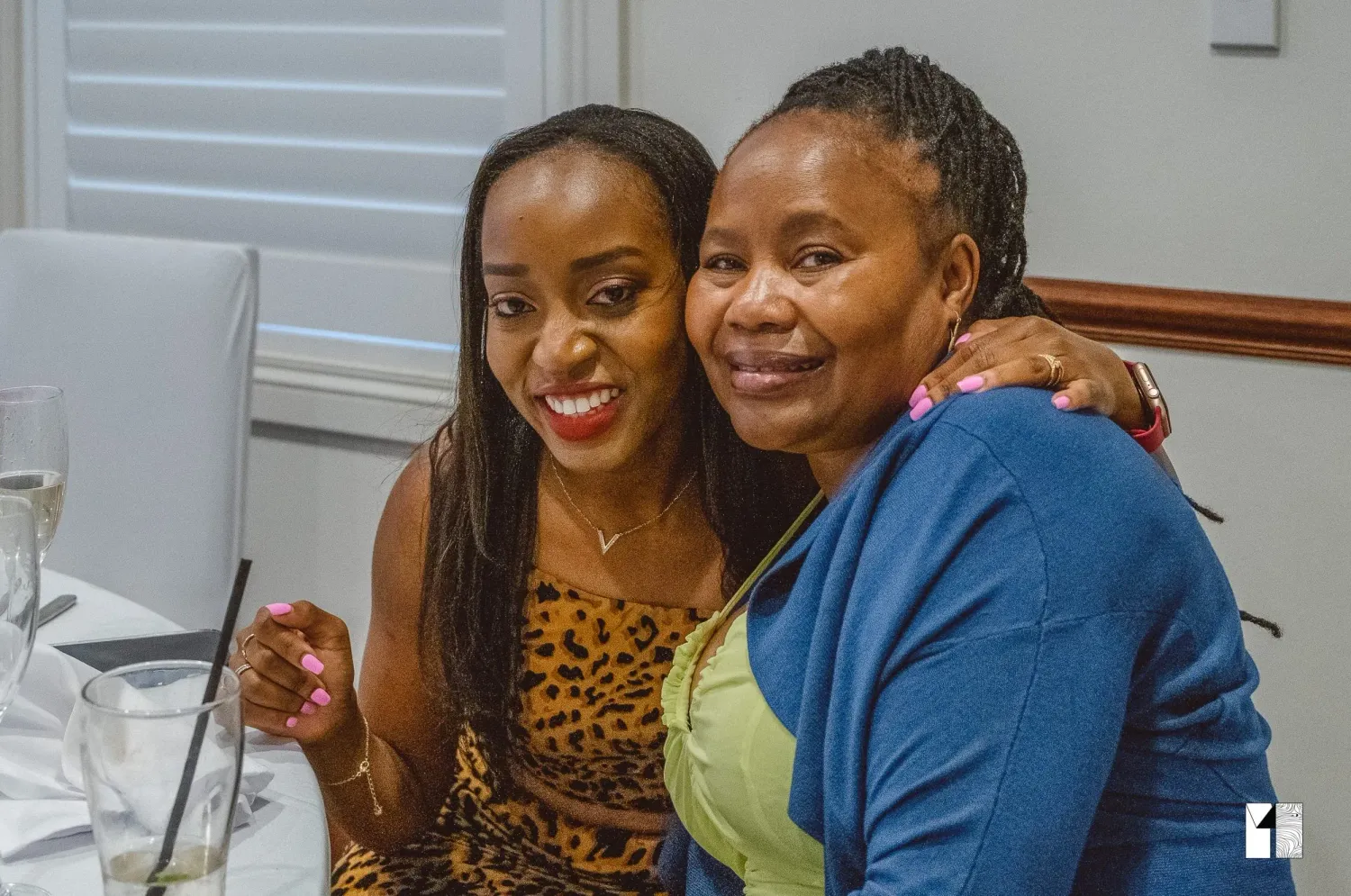 Two women smiling and embracing at an indoor wedding reception dinner celebration.