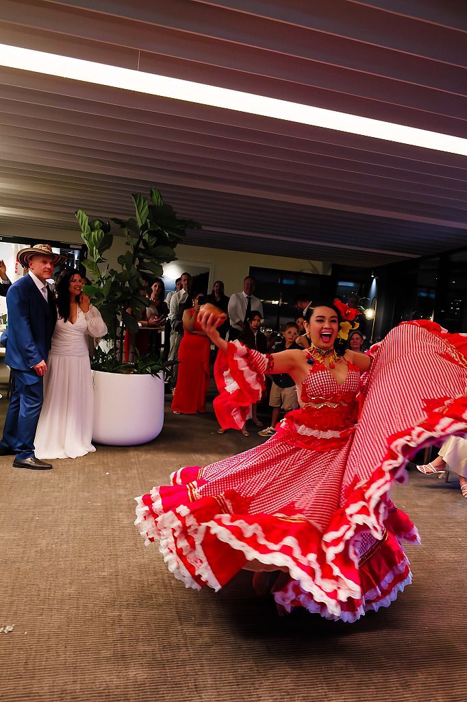 A dancer in a red and pink ruffled skirt twirls at a wedding reception while the bride and groom watch in the background.