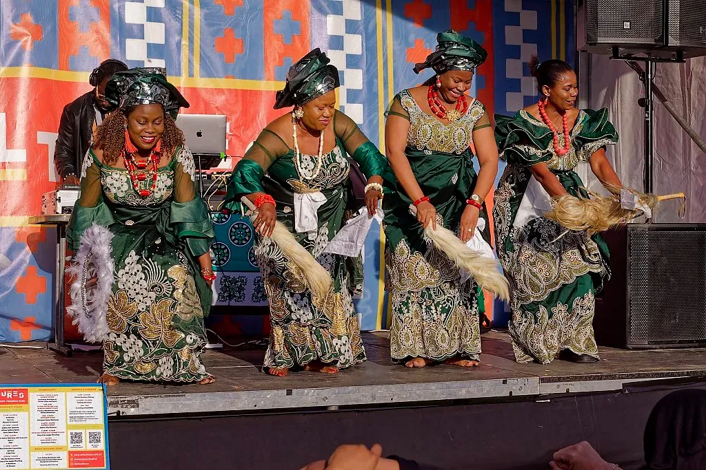 Four women in elegant green and gold traditional African attire dance together on stage.