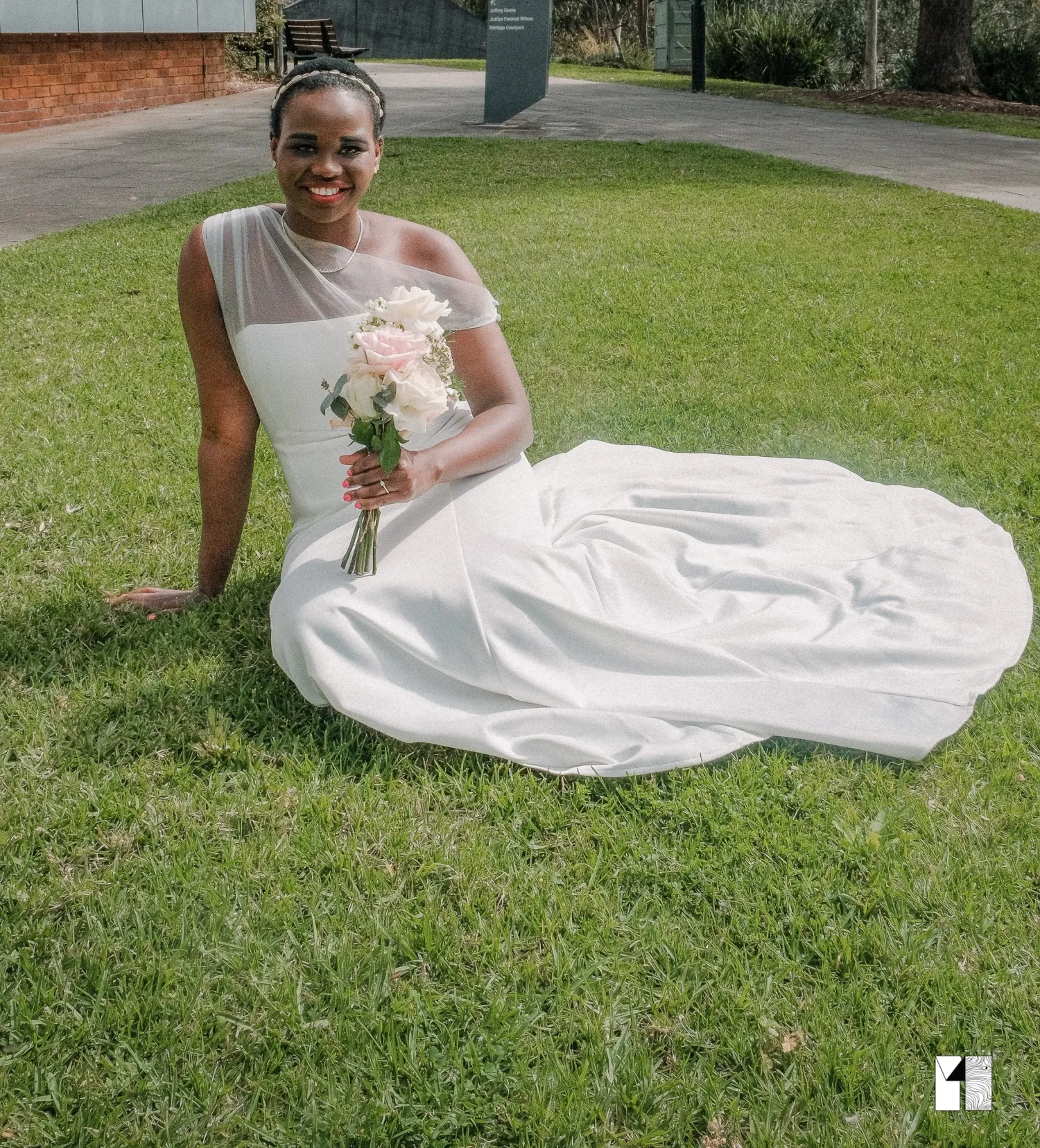 Smiling bride in white gown sitting on grass holding a pink rose bouquet.