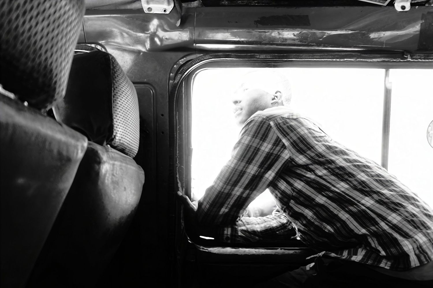 Black and white photo of a man in a plaid shirt leaning out of an open vehicle window, gazing outside from a worn bus interior.