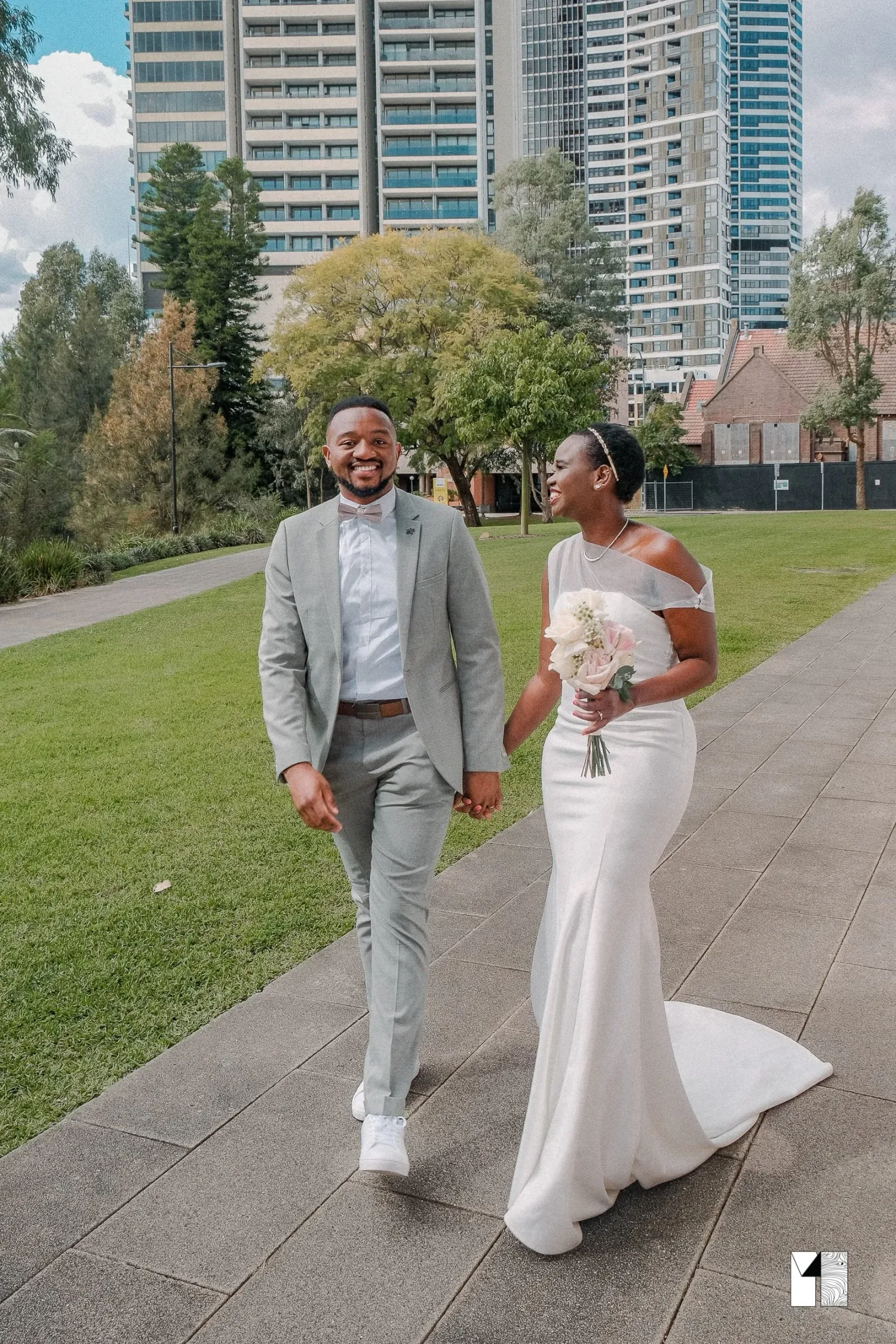 Bride and groom walking together in an urban park, bride in white gown, groom in gray suit.