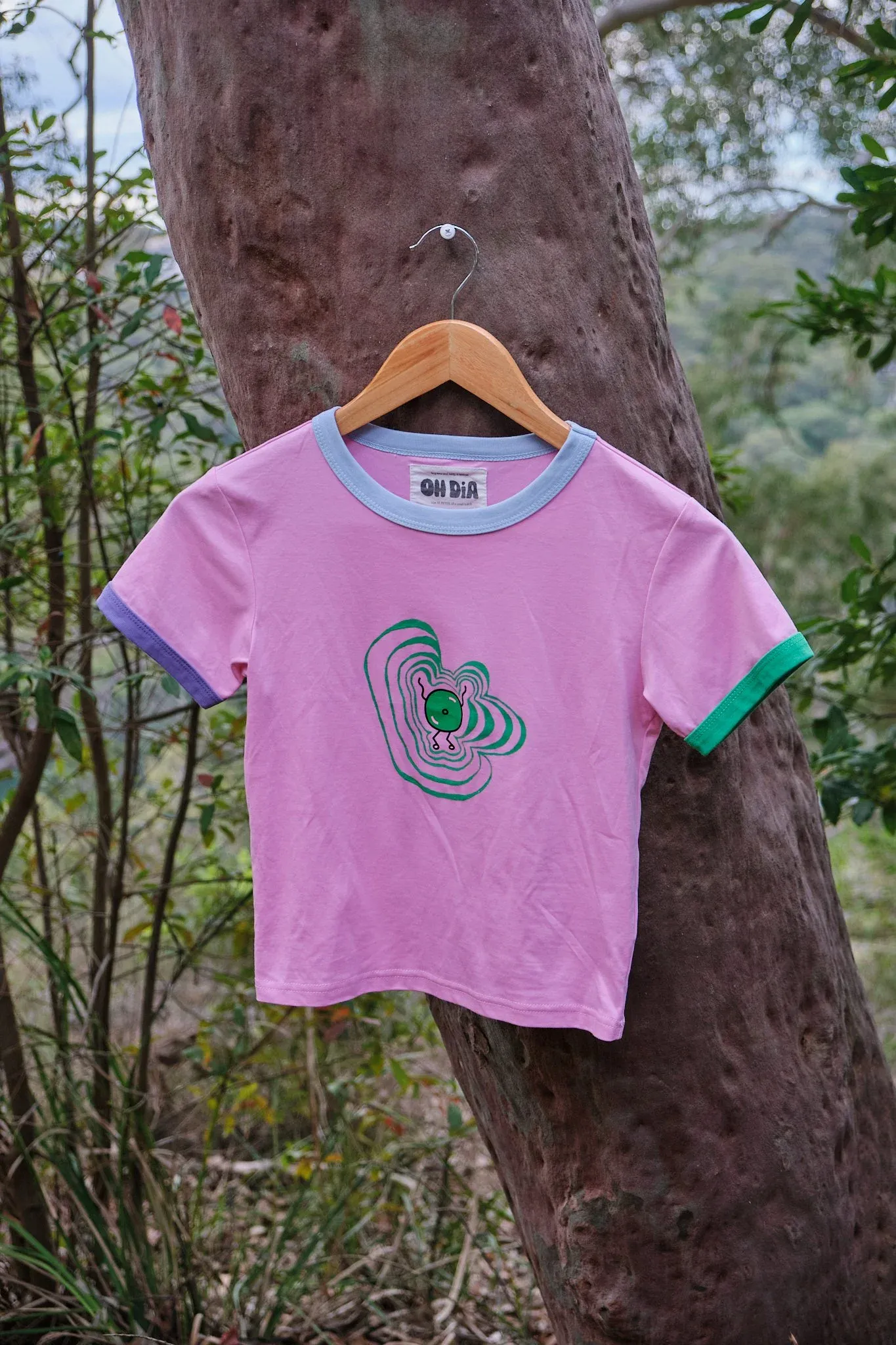 Pink crop ringer tee with teal abstract print on wooden hanger against tree.