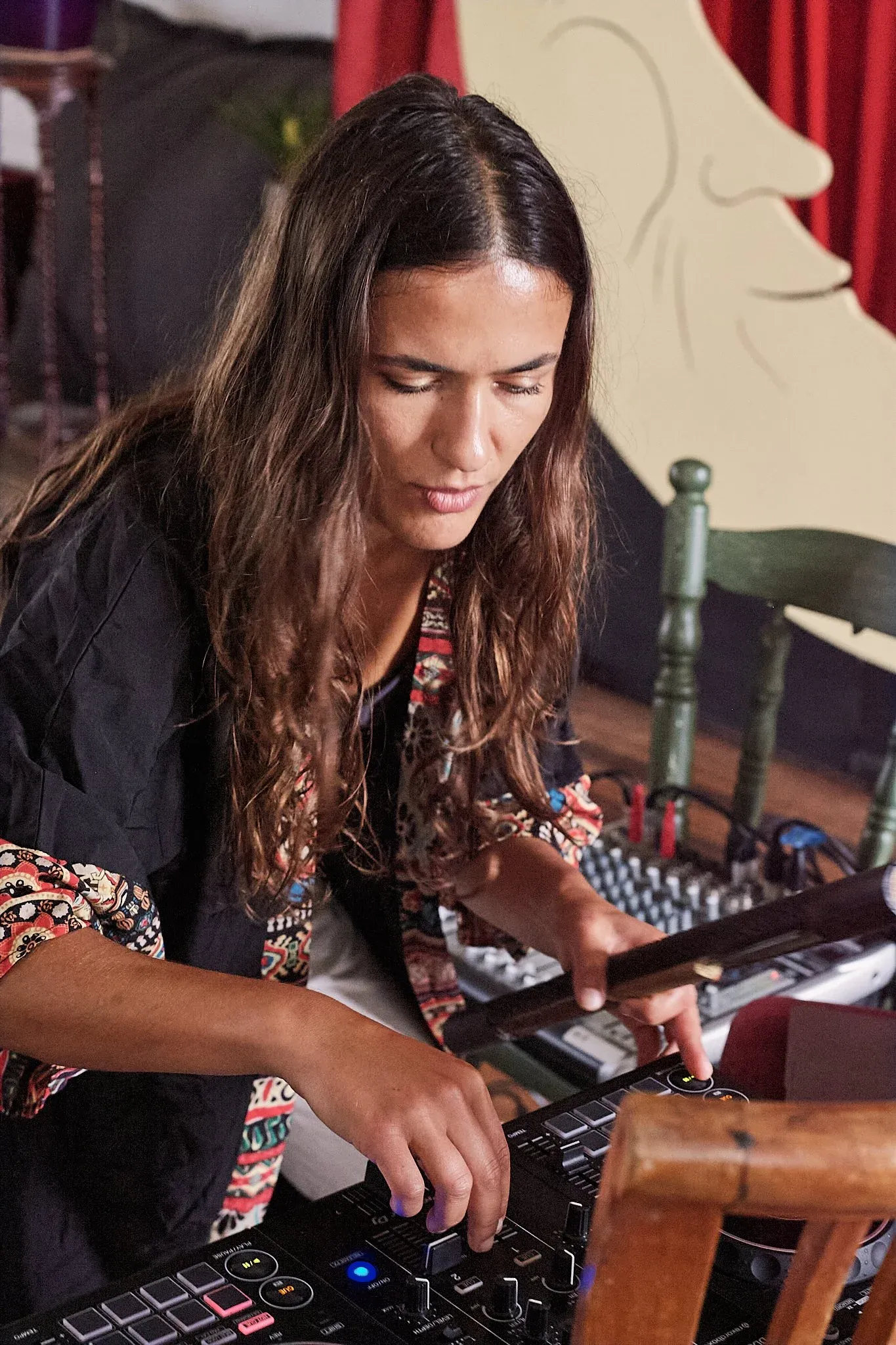 Female musician with long hair focuses on mixing equipment in a colorfully decorated performance space.
