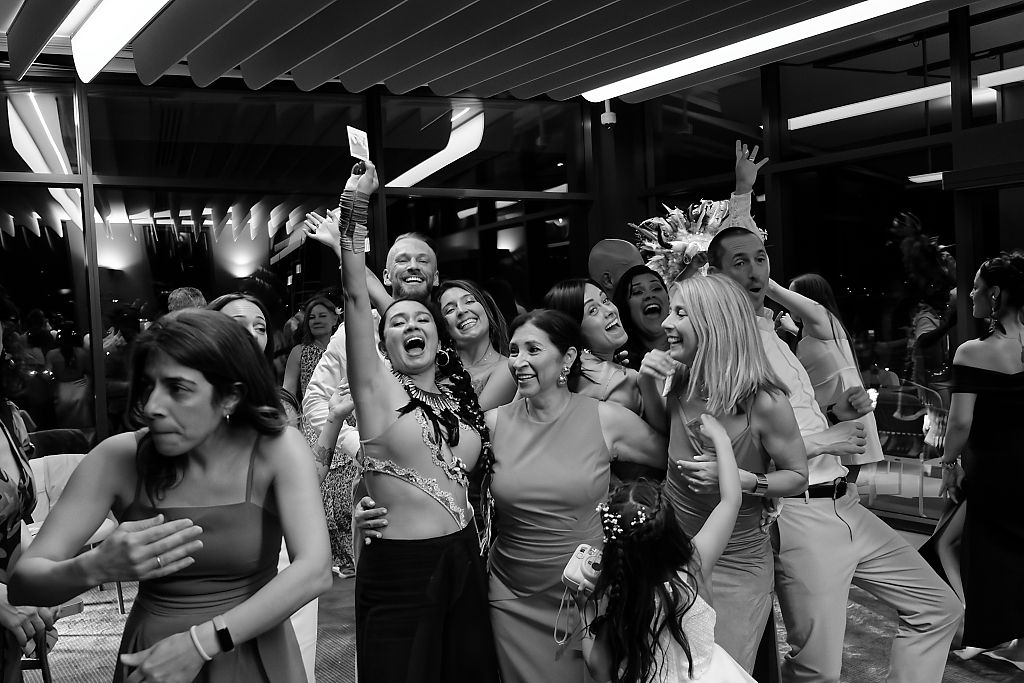 Black and white photo of cheering wedding guests with raised arms laughing and celebrating together on a lively indoor dance floor.