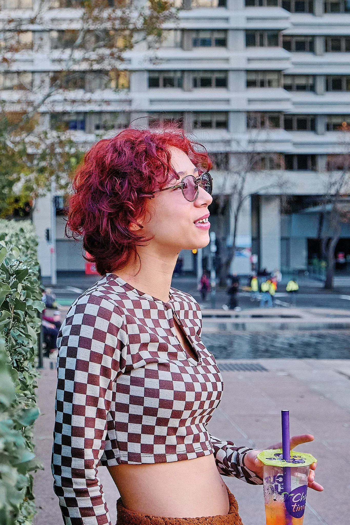Red-haired woman in checkered crop top holding bubble tea drink in urban city setting.