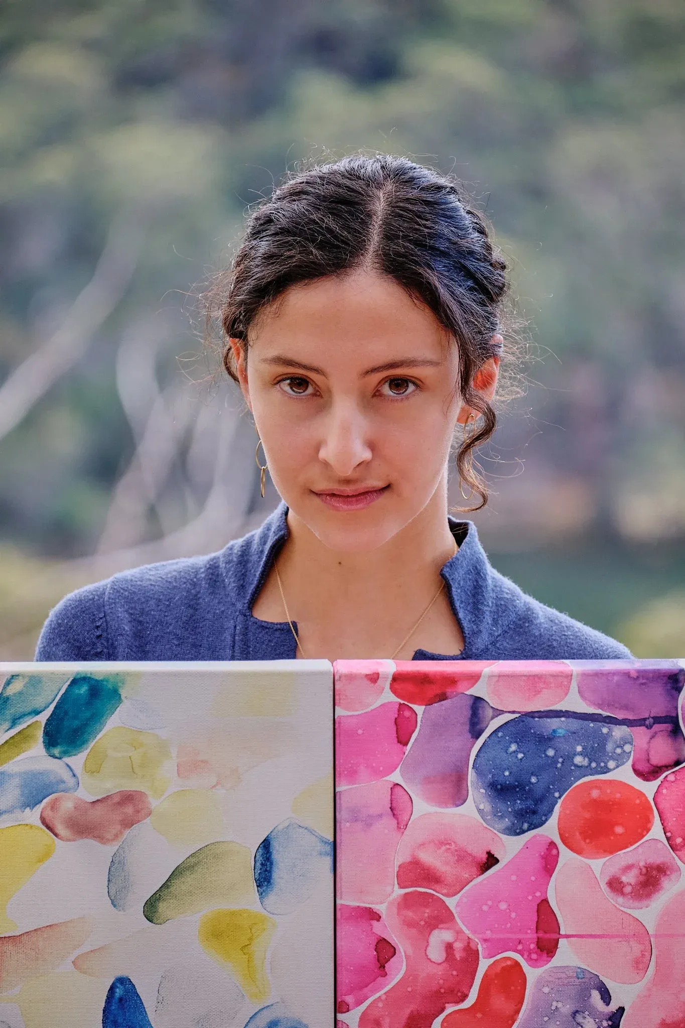Young woman holding colorful watercolor paintings outdoors with blurred green background.