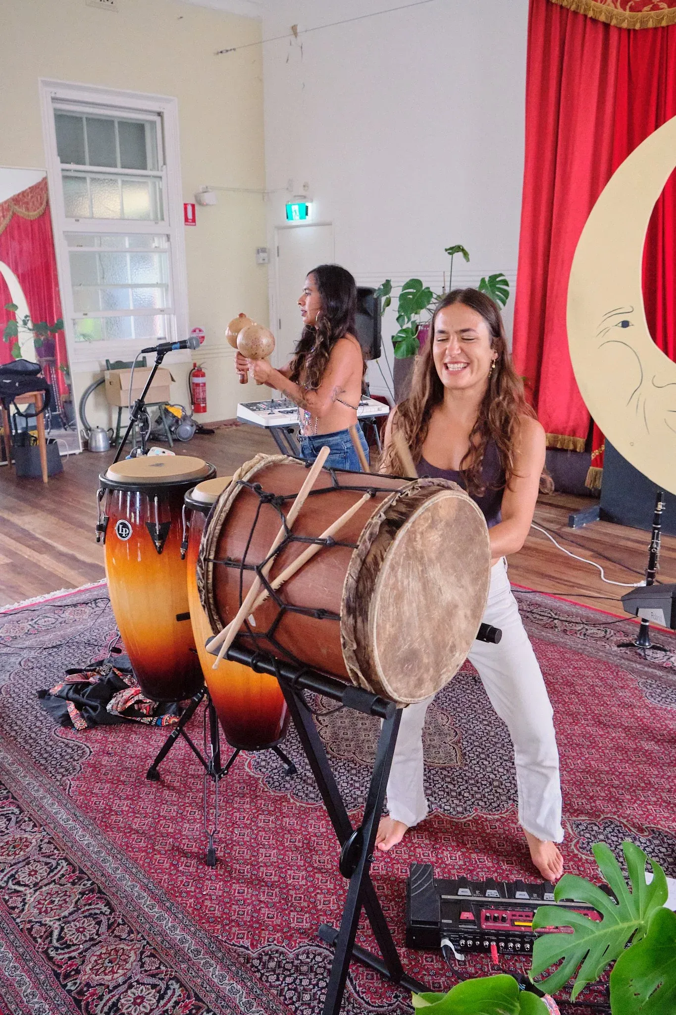 Woman energetically playing a large frame drum on a stand in a vibrant music studio with red curtains.