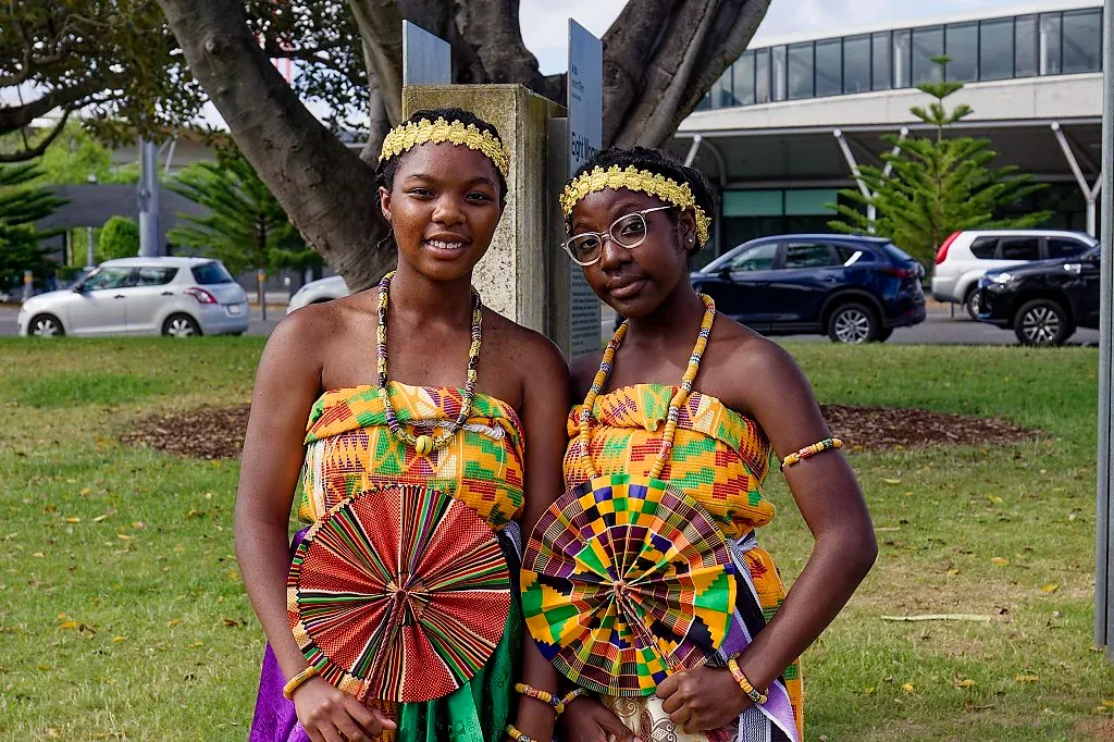 Two young women in matching kente cloth African traditional dresses holding decorative fans outdoors.
