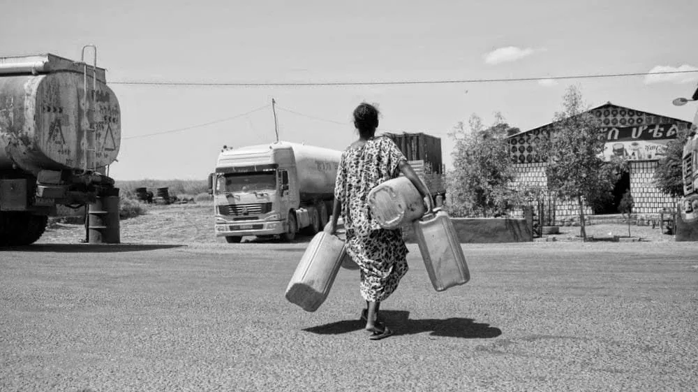 Black and white photo of a woman carrying large water jugs walking toward a tanker truck on a rural road.