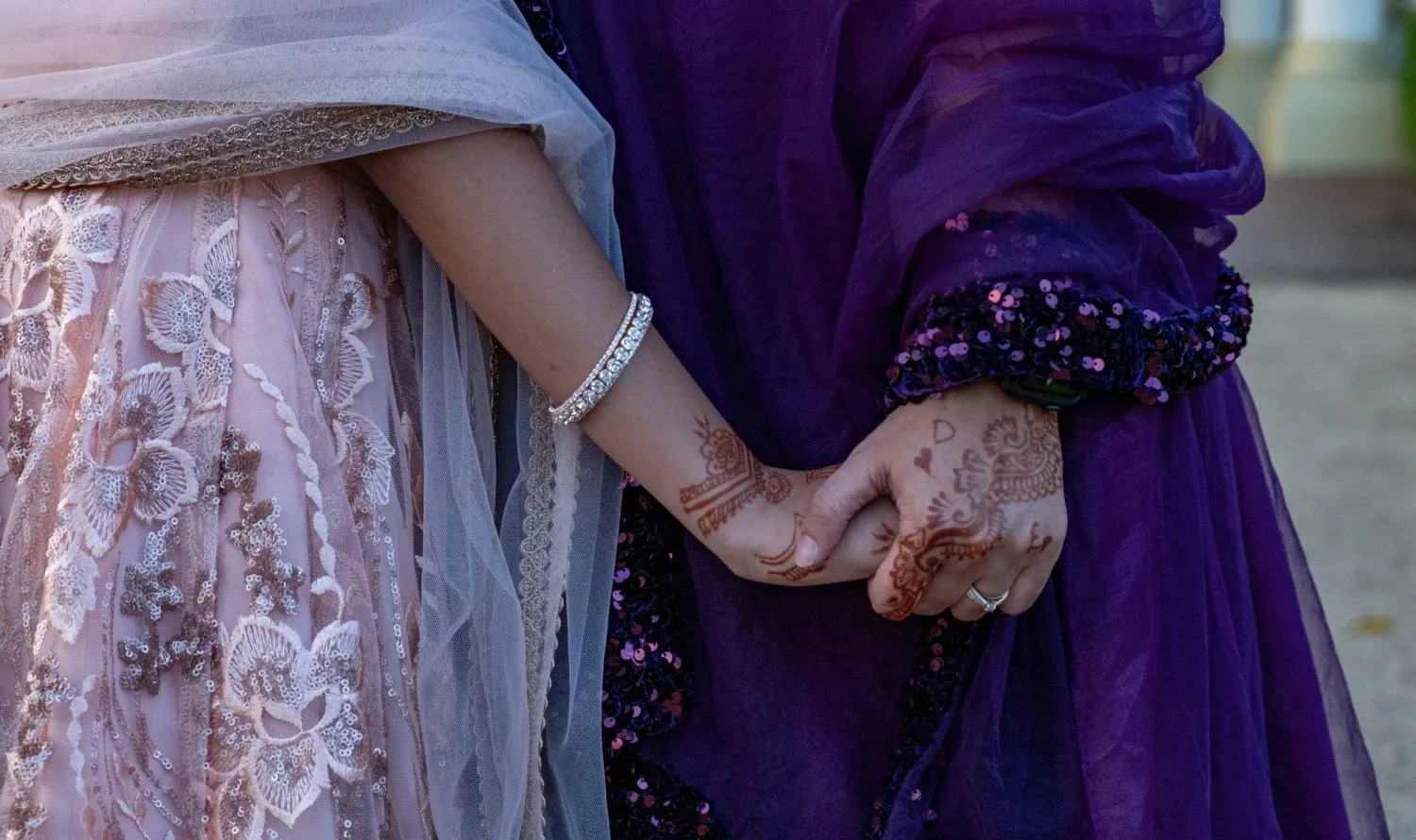 Two women in elegant South Asian attire hold hands, showcasing henna and pearl bracelet details.