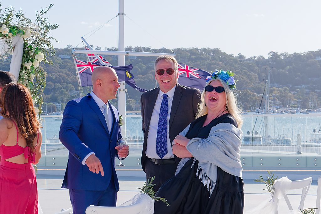 Wedding guests in formal attire laugh together on a waterfront deck with Union Jack flags, floral decor and a marina behind them.