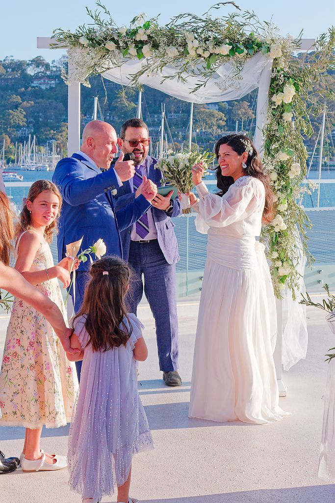 Bride in a white dress holds a bouquet beneath a floral arch as the celebrant and flower girls look on by the water.