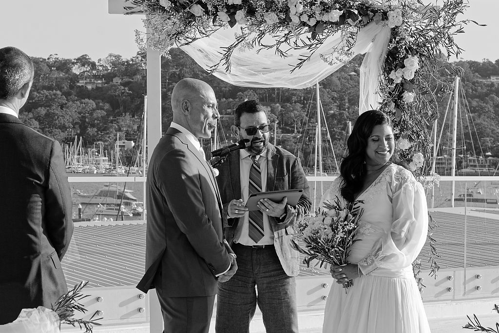 Bride and groom exchange vows under a flower-draped wedding arch as the officiant reads, with a waterfront marina behind them.