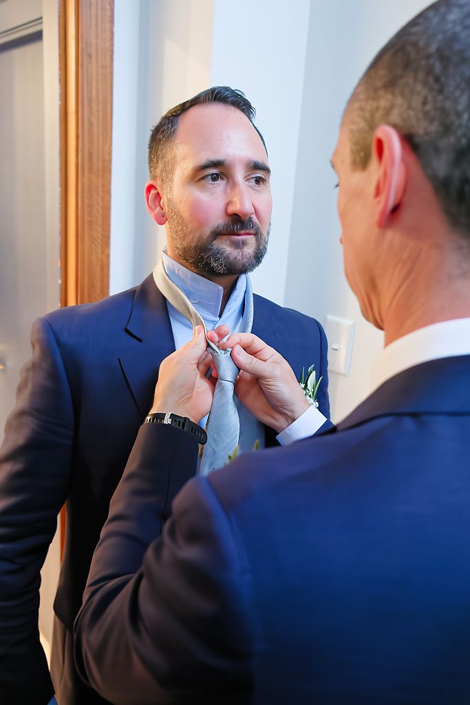 Groom in a navy suit looks on as another man in a matching jacket adjusts his gray patterned tie before the wedding.