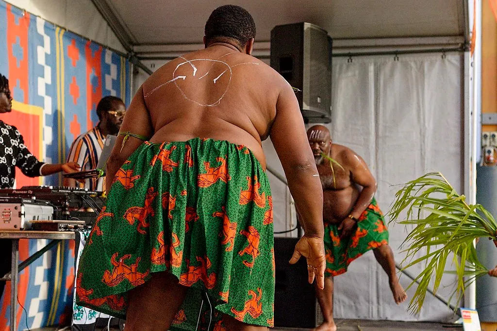 Two shirtless men in vibrant green and orange floral skirts dancing at cultural festival.