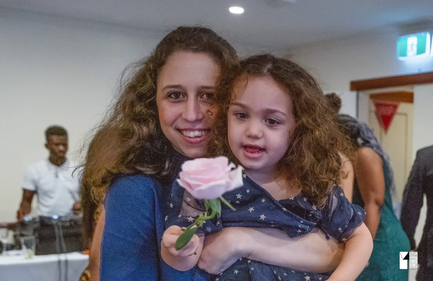 Smiling woman holding a young girl with a pink rose at a wedding reception.