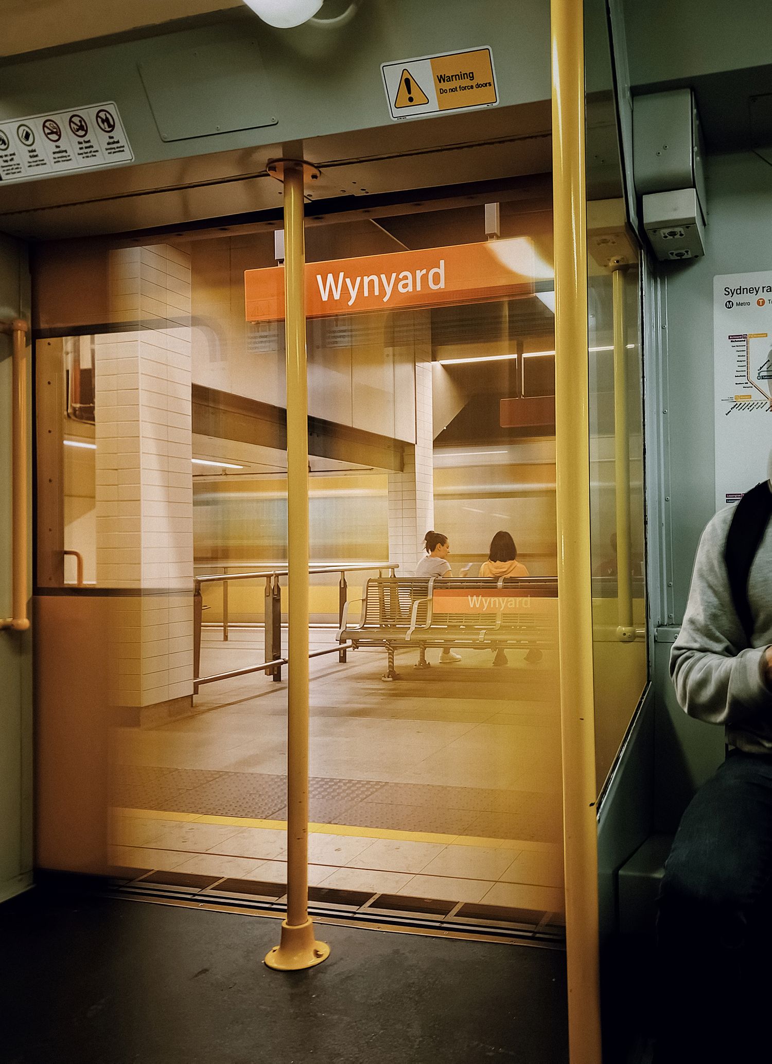 Train doorway view of Wynyard station platform in Sydney, with two people seated on a bench and a passenger in the foreground.