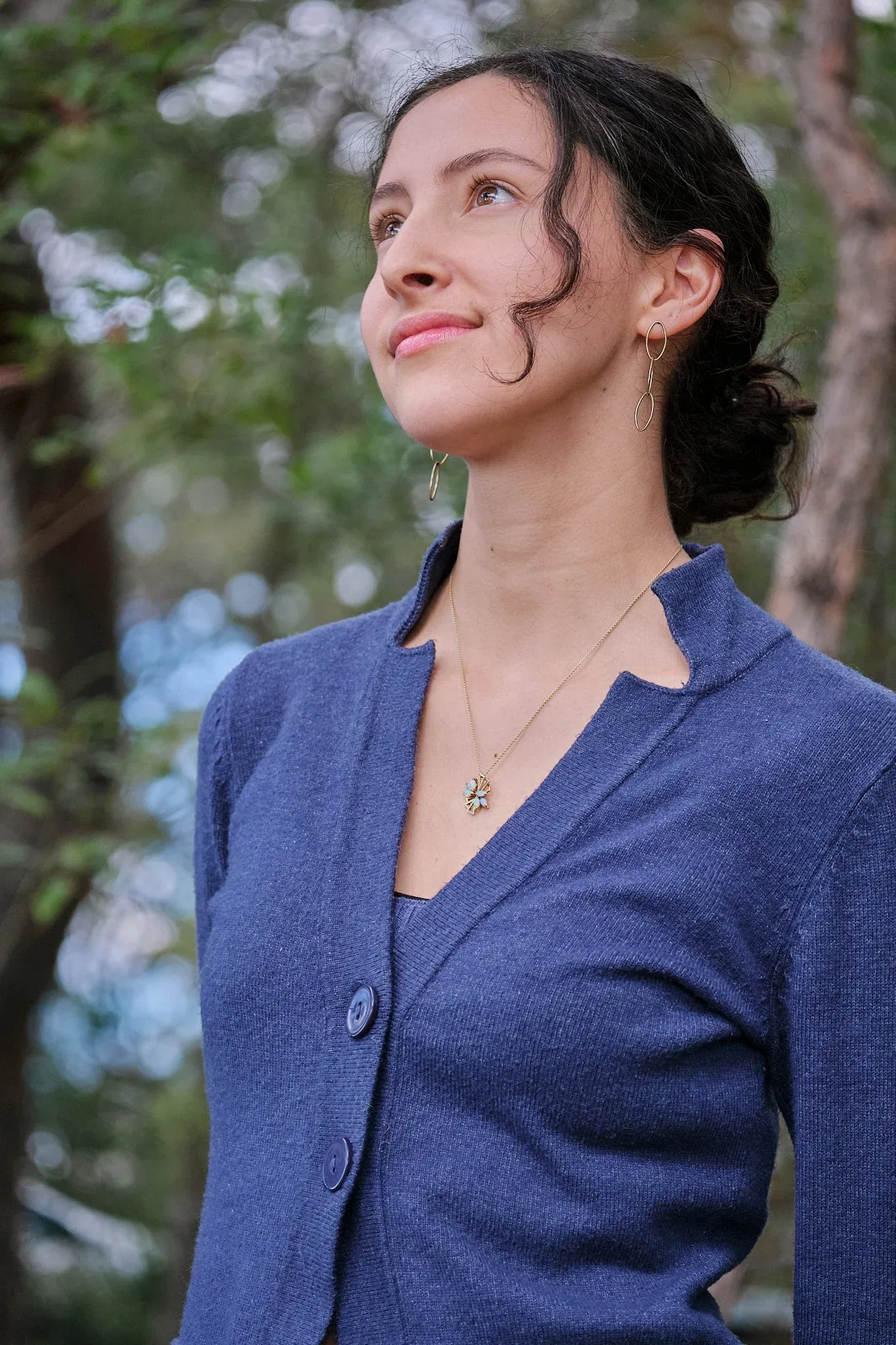 Woman in blue cardigan with gold necklace and earrings gazing upward outdoors among trees.