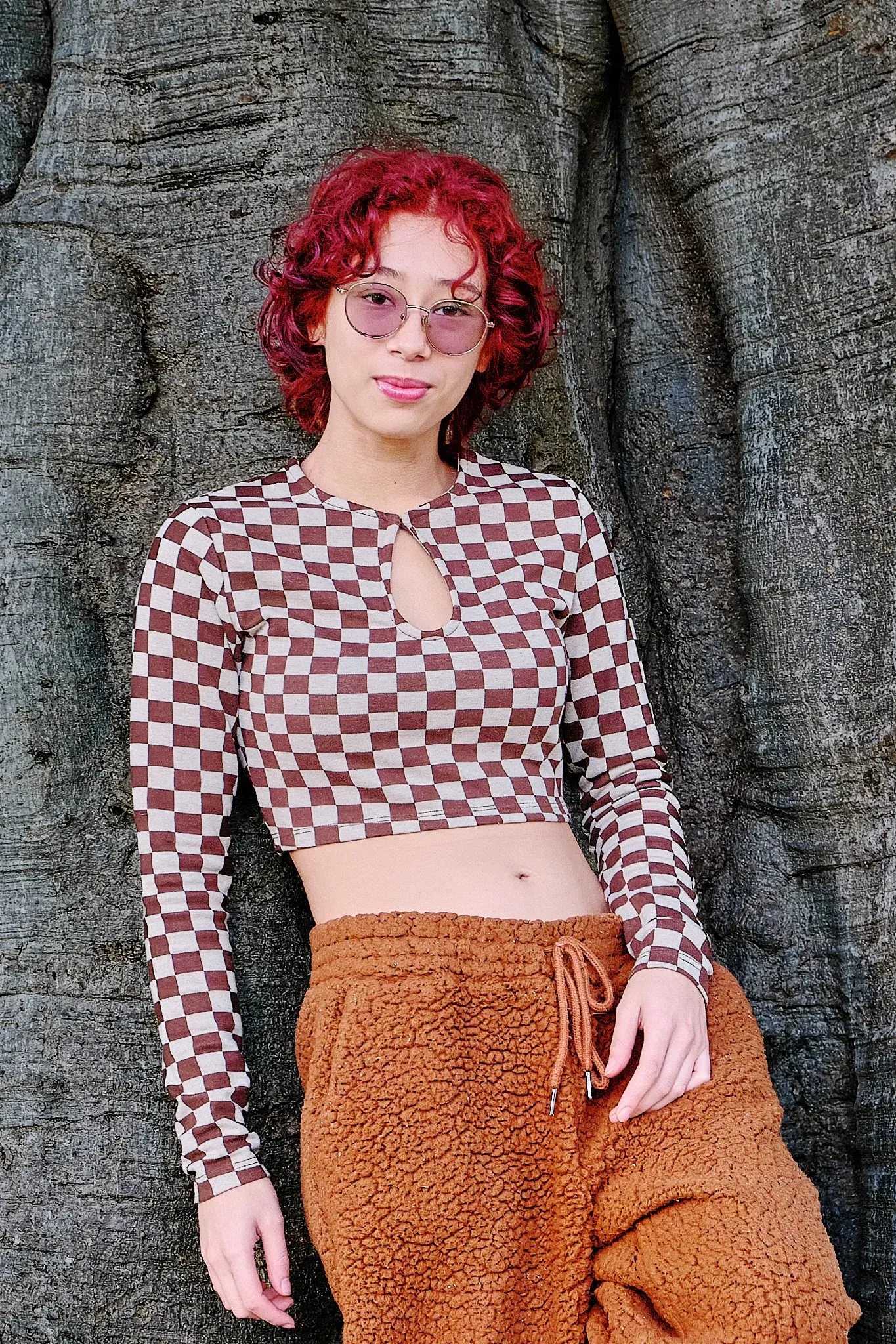 Young woman with red curly hair wearing checkered crop top and sunglasses leaning against tree bark.