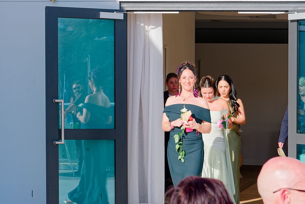 Bridesmaids in green dresses carrying bouquets walk toward an outdoor wedding ceremony as guests look on.