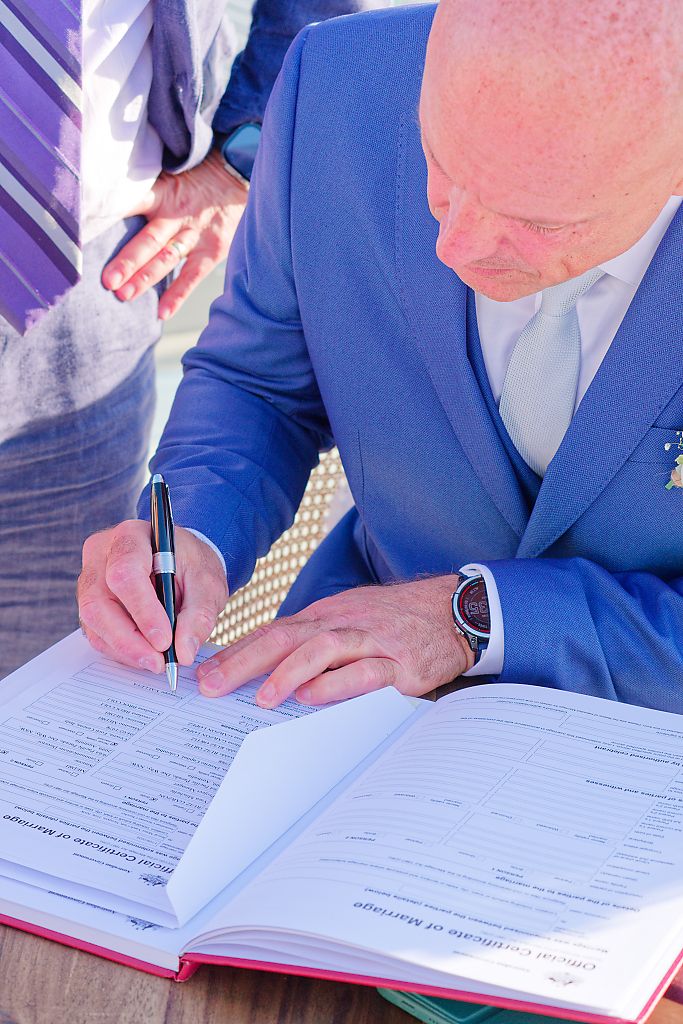 Groom in a blue suit signs the official certificate of marriage with a black pen at a wedding ceremony.