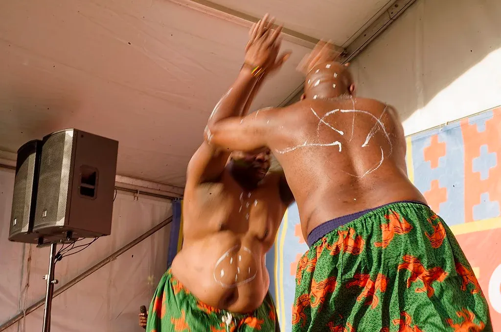Two shirtless male dancers performing traditional dance on outdoor festival stage.