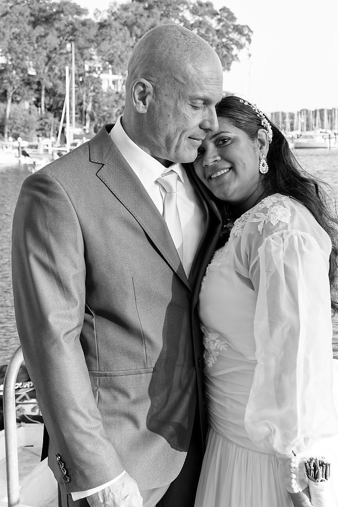Black and white wedding portrait of a bride in a lace dress and veil nestled against her groom in a suit beside a marina.