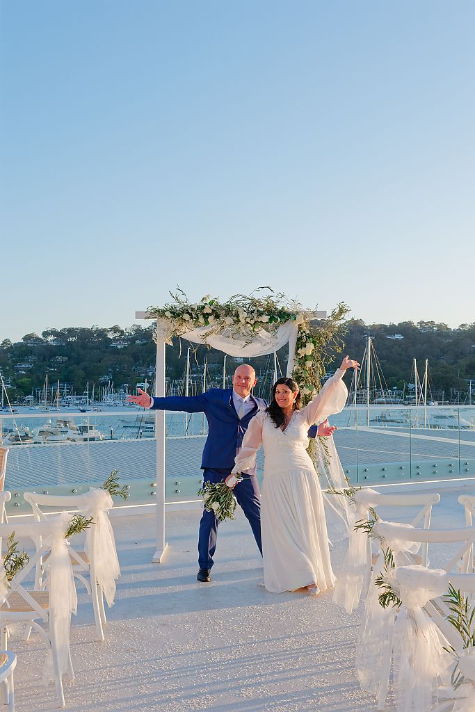 Bride in a white gown and groom in a blue suit celebrate with raised arms under a floral wedding arch on a sunny waterfront terrace.