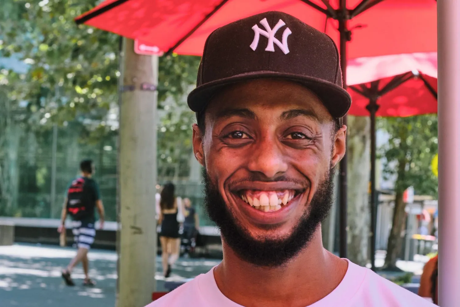 Smiling Black man wearing brown NY Yankees cap laughing outdoors under red umbrella in park.