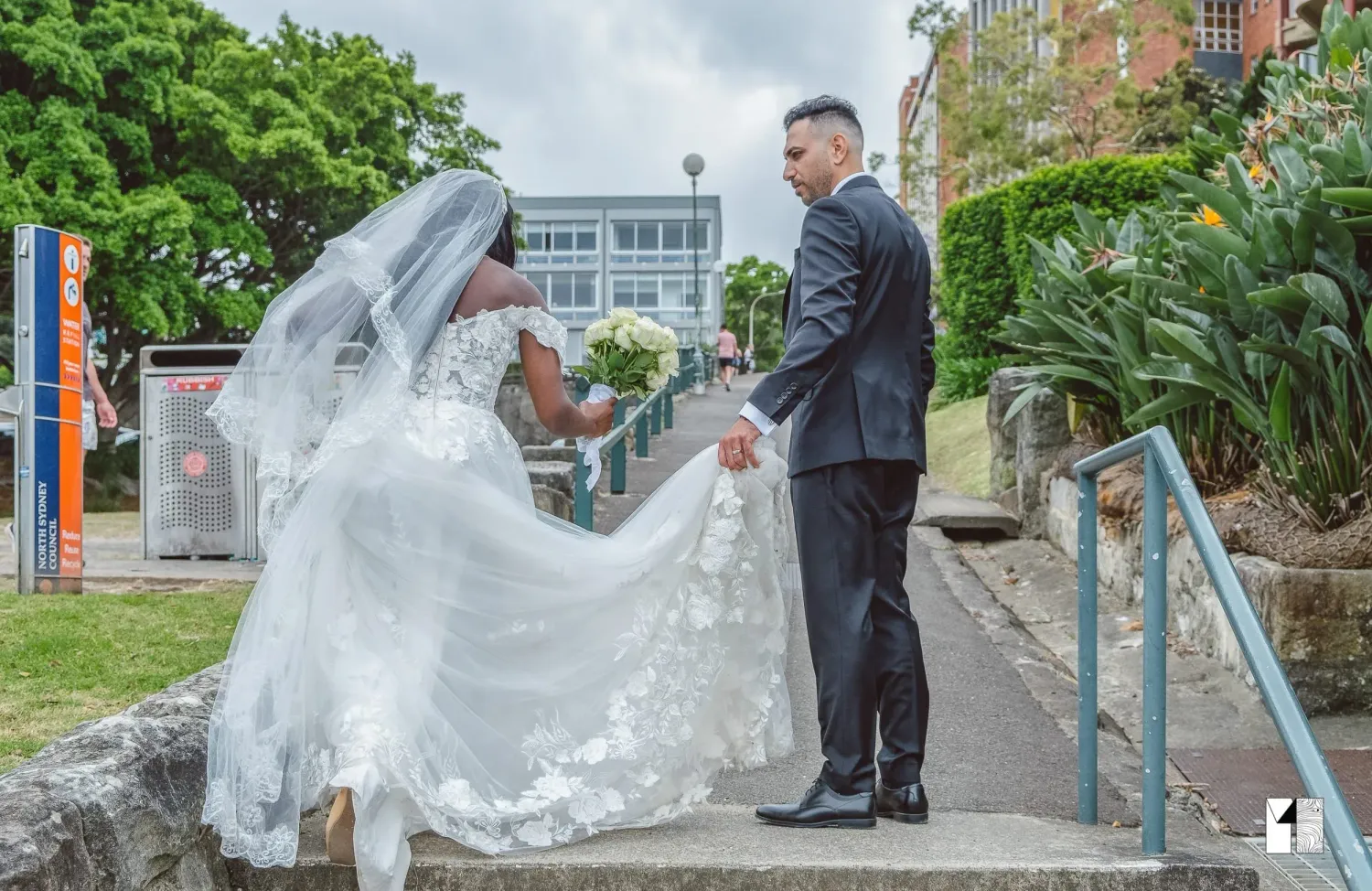 Bride in elegant lace gown and groom hold hands on steps during outdoor wedding.