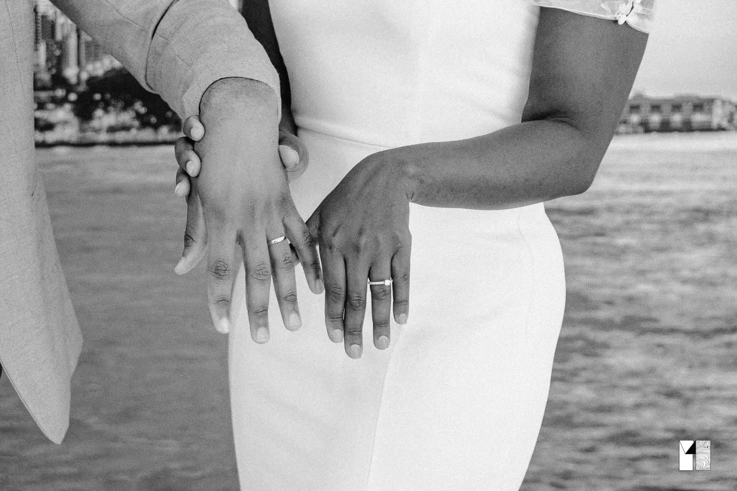 Close-up black and white photo of newlywed couple showing wedding rings near waterfront bridge.