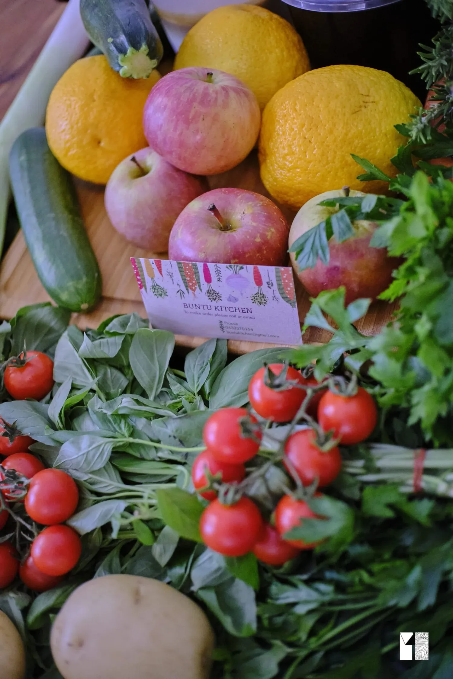 Fresh produce display with apples, oranges, cucumbers, cherry tomatoes, and herbs on a wooden board.